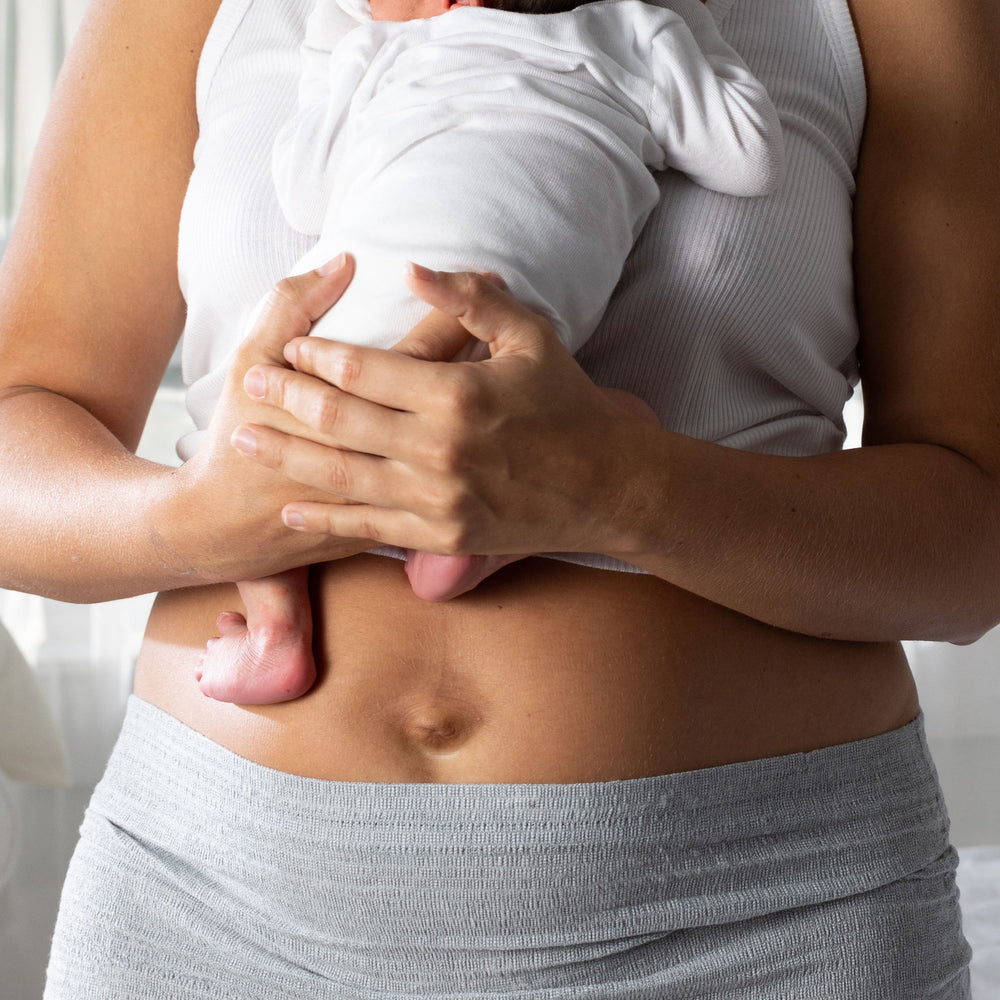 Woman holding a baby wrapped in a white blanket, wearing a white tank top and gray shorts.